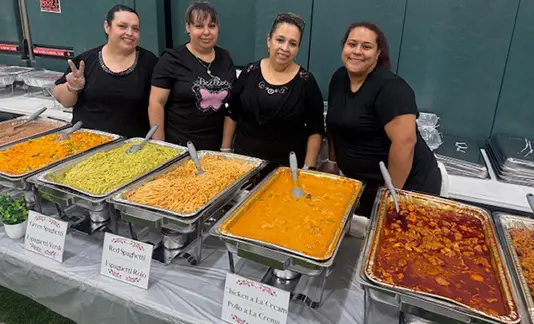 Four women standing behind a table with trays of various foods.